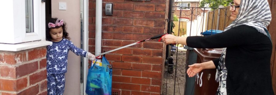 A woman hands a plastic bag of jigsaws and crafts - using a litter picker - to a child stood in a doorway, wearing plastic gloves