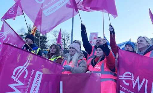 Striking postal workers gather on a picket line waving flags from the CWU union