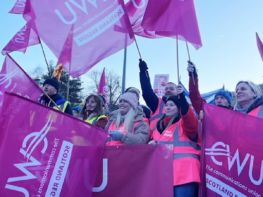 Striking postal workers gather on a picket line waving flags from the CWU union