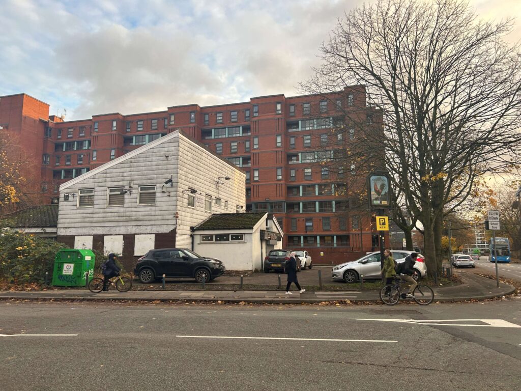 A photo of the abandoned Gamecock Pub, a dirty white building with a large red brick building behind it. The windows are shuttered, and a large sign with a cockerel stands outside.