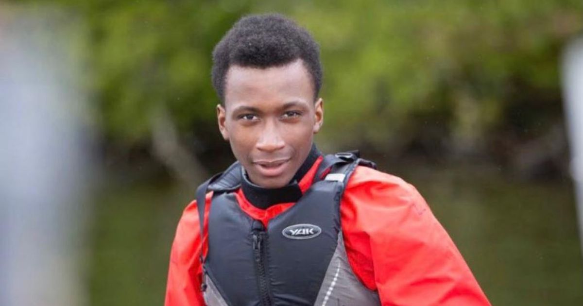 Headshot of Ronaldo Johnson. He is a Black teenage male wearing a red waterproof jacket and black life jacket, staring directly into the camera and smiling. Behind him is a river and green foliage. He is in a canoe, but the canoe has been cropped out.