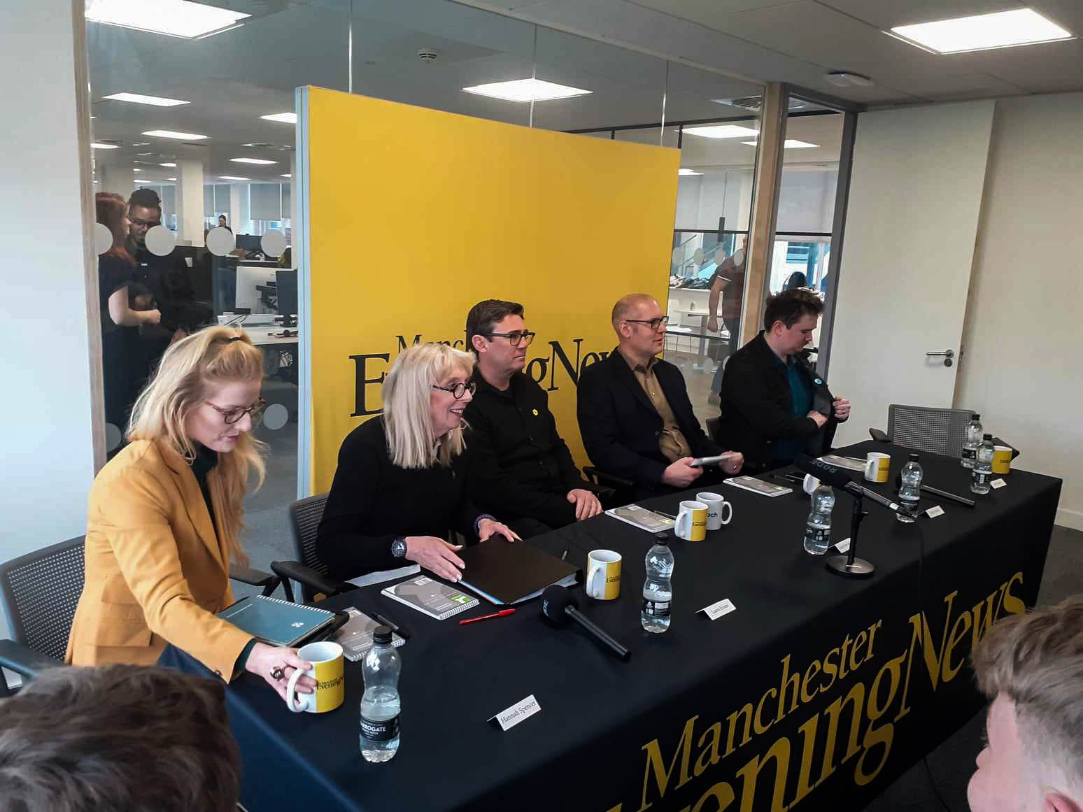 Mayoral candidates sitting at a table in the Manchester Evening News offices. From left to right: Hannah Spencer, Laura Evans, Andy Burnham, Dan Barker, and Jake Austin. Each candidate has a notebook, a mug, and a bottle of water. There is a yellow backdrop with the Manchester Evening News logo.