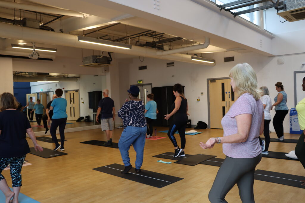 Large group of people stood in fitness studio on yoga mats as they do a gentle circuit class.