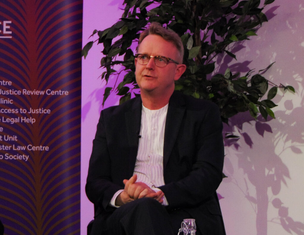 Pete Weatherby, a white man in a black blazer and white shirt, sits on a chair mid-speech with his hands clasped.