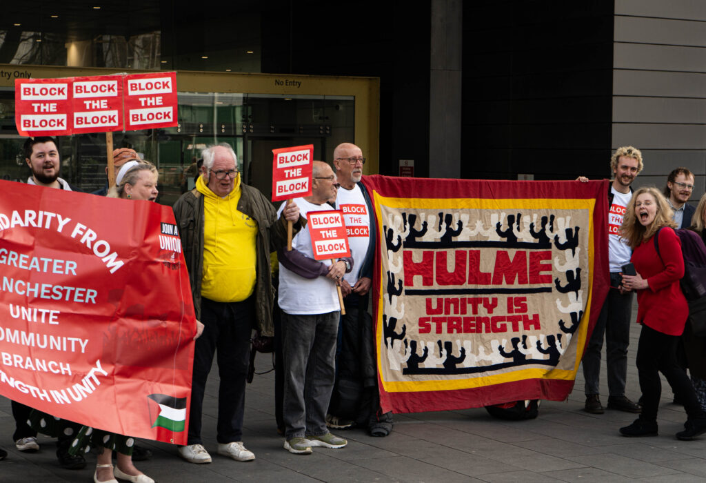 A group of people stand on the pavement outside Manchester County Court. They hold red signs that say 'Block the Block', as well as a large fabric sign that says 'Hulme: Unity is strength'