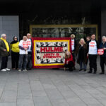 A group of people stood on the pavement outside Manchester County Court. They hold small cardboard signs that say "Block the Block" and a large cloth sign that says "Hulme: Unity is strength"