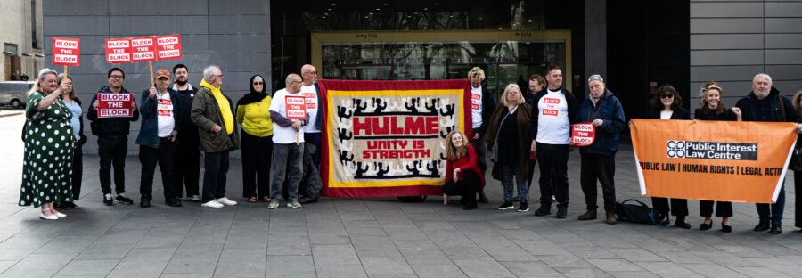 A group of people stood on the pavement outside Manchester County Court. They hold small cardboard signs that say "Block the Block" and a large cloth sign that says "Hulme: Unity is strength"