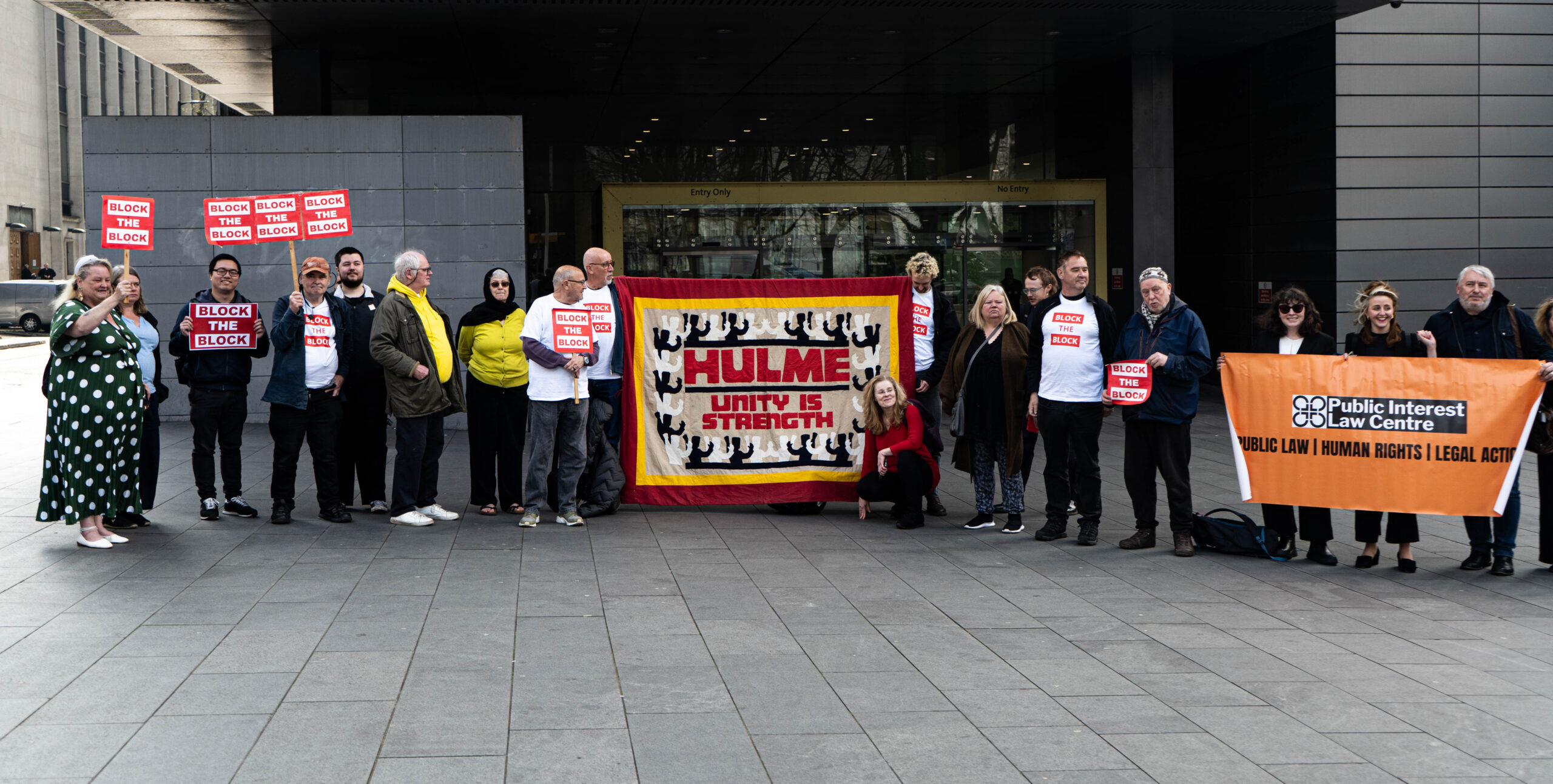 A group of people stood on the pavement outside Manchester County Court. They hold small cardboard signs that say "Block the Block" and a large cloth sign that says "Hulme: Unity is strength"