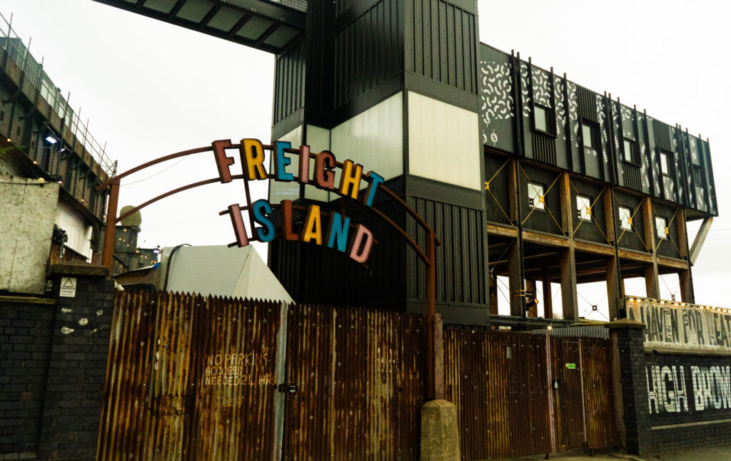 the front of Freight Island. The rusted gates are shuttered, and a large sign that says 'FREIGHT ISLAND' arcs above it. The rest of the building is black and white and built on pillars in the background. The area is closed, and the sky is grey.