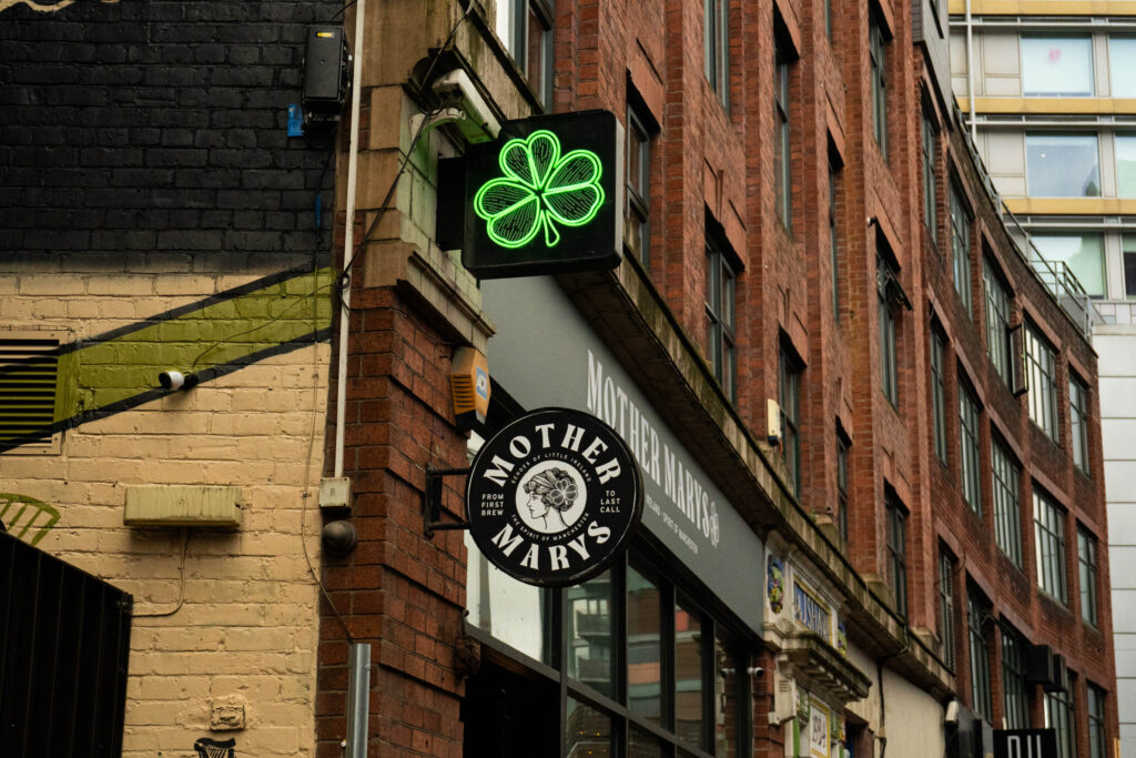 The two signs sticking out of Mother Mary's bar. the top sign is of a glowing neon green shamrock. Beneath is a circular sign in black and white, which says 'MOTHER MARY''S' around the logo: a profile illustration of a woman with a shamrock over her ears. The rest of the building is visible out of focus as the street curves into the distance.