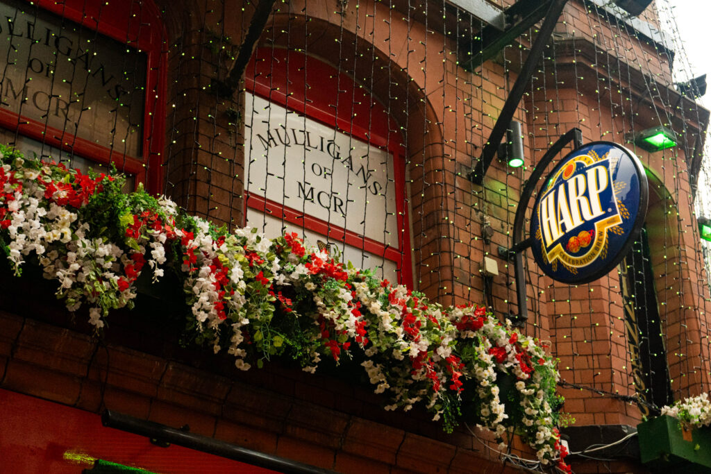 A window sign above the door of Mulligans of Deansgate. It says 'MULLIGANS OF MCR' in the window, which has a red frame. A round blue sign that says 'HARP CELEBRATION BREW' sticks out of the side of the building. Both signs are behind strings of fairy lights, and above a long tray of fake red and white flowers.