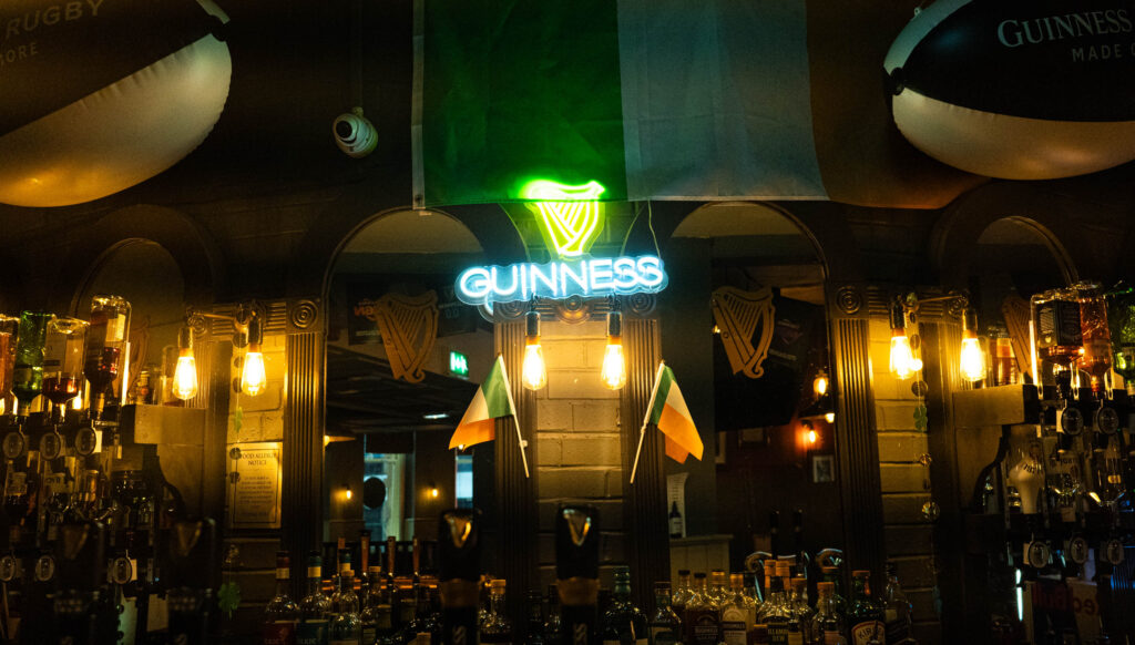A display behind O'Sheas' bar. A neon sign that reads 'GUINNESS' in white sits below a neon green Guinness logo. Two small Irish flags hang beneath, with a larger flag above. The space is dark, and lit with warm lightbulbs strung about. Various bottle of alcohol are visible behind the bar.