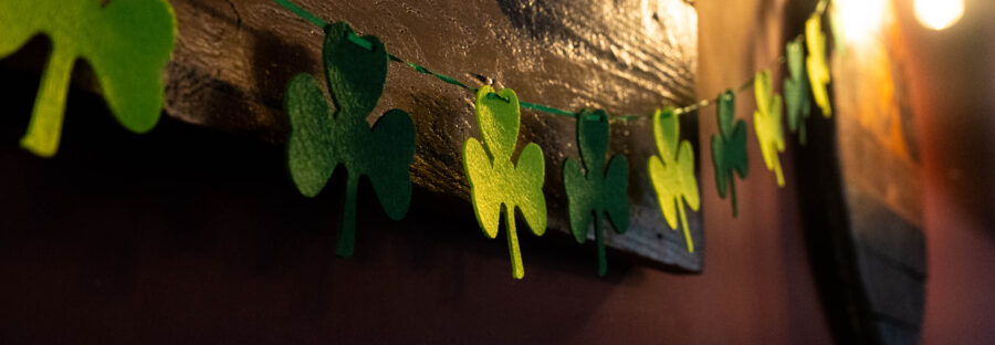 A string of green, clover shaped bunting made of felt. It is strung across a wooden wall, and there is a bright, warm lightbulb in the top right corner.