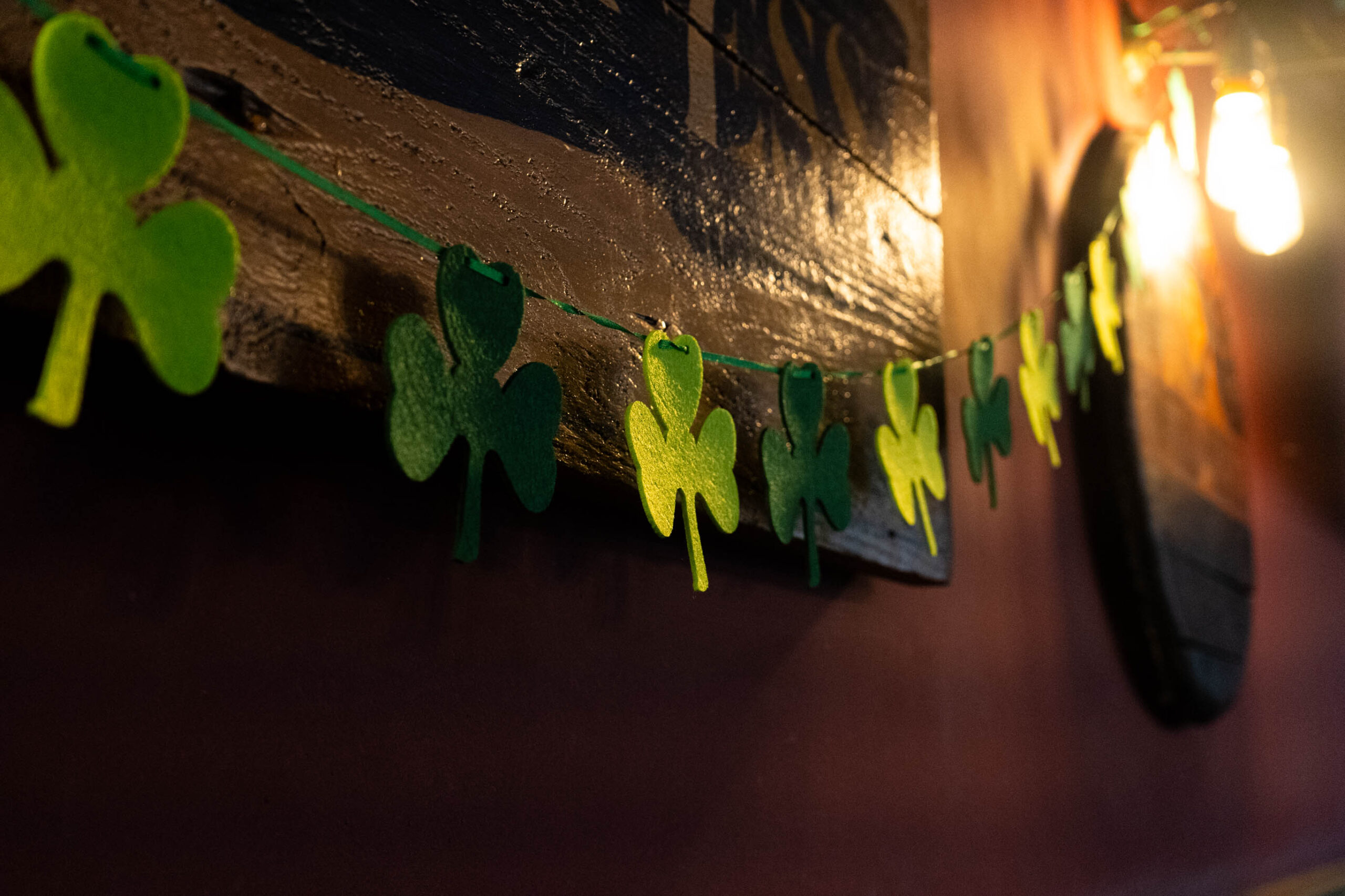 A string of green, clover shaped bunting made of felt. It is strung across a wooden wall, and there is a bright, warm lightbulb in the top right corner.