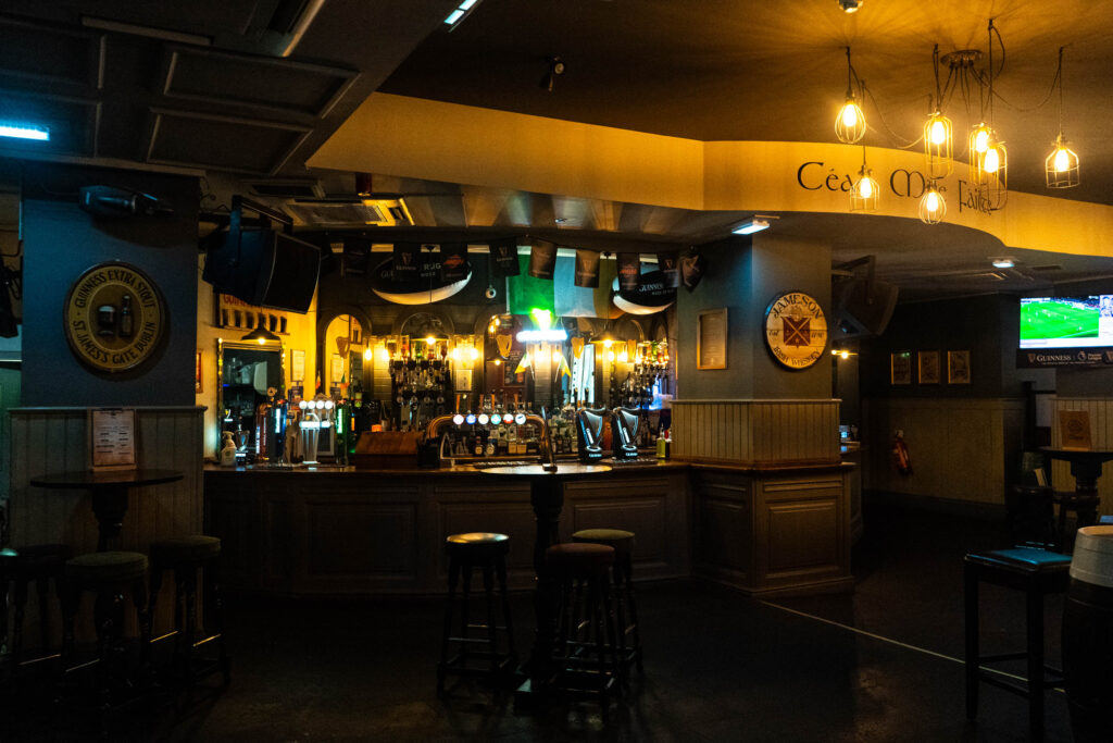The bar in O'Sheas. It is dark and lit by warm lightbulbs strung about. There are dark oak tables visible, and behind the bar is cluttered with lights, alcohol, and a large Irish flag hanging above.