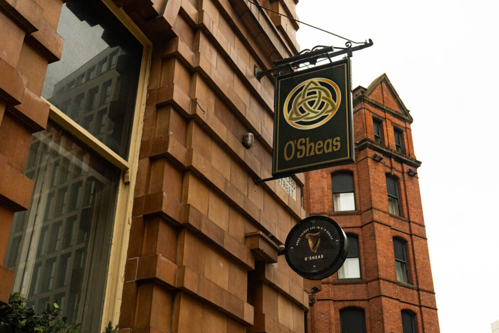 the signs sticking out of O'Sheas Irish Bar. It is a black sign which says 'O'SHEAS' underneath the logo, a gold and green Celtic knot. Underneath is a smaller sign advertising Guinness. the signs stick out of a brown, Victorian brick wall.