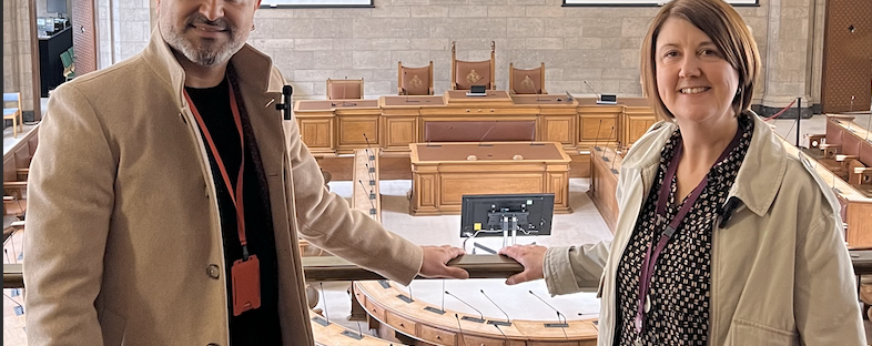 Faraz Arian stands on the left of the image in a dark shirt, and cream overcoat and hat. He is smiling at the camera with his arm stretched onto a bannister. On the right of the image, Joanna Midgley wears a cream jacket and purple shirt. She is a white woman with a brown bob, and she is also smiling at the amera with an arm on the bannister. They are stood on a balcony, and below them is a well-lit council chamber