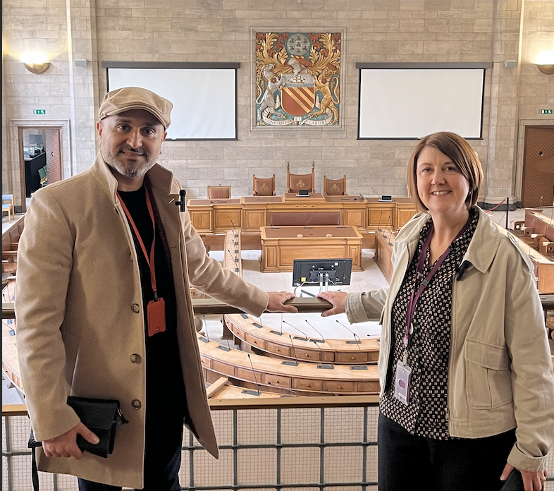 Faraz Arian stands on the left of the image in a dark shirt, and cream overcoat and hat. He is smiling at the camera with his arm stretched onto a bannister. On the right of the image, Joanna Midgley wears a cream jacket and purple shirt. She is a white woman with a brown bob, and she is also smiling at the amera with an arm on the bannister. They are stood on a balcony, and below them is a well-lit council chamber