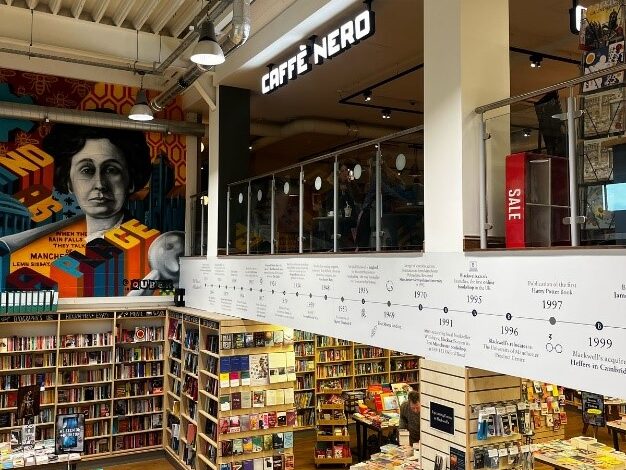 The interior of Blackwells bookshop, taken from a staircase. The camera is pointed between two floors: on the ground floor is a large room filled with bookshelves; on the first floor is a Caffè Nero, separated by a glass barrier. On the floor in between is a timeline of the bookshop's history. On the left of the image is a large red, blue, and yellow Brutalist mural of a woman painted above the bookshelves. 