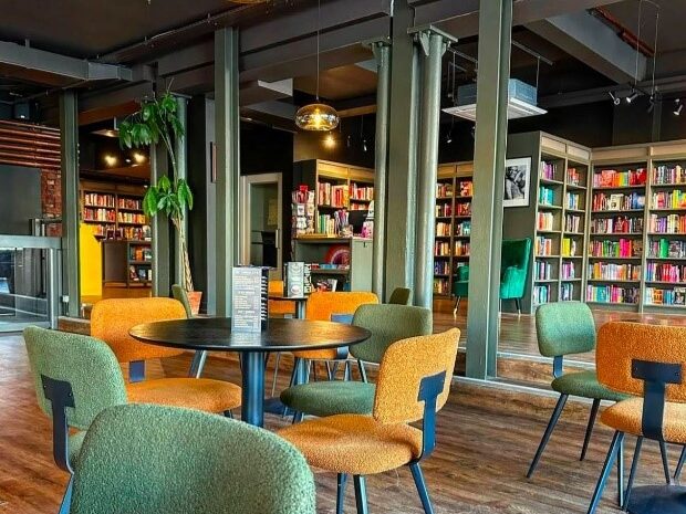 The interior of Queer Lit bookshop. Several round, black tables with yellow and green chairs are visible in the foreground. In the background behind some green painted pillars is a room filled with bookshelves.