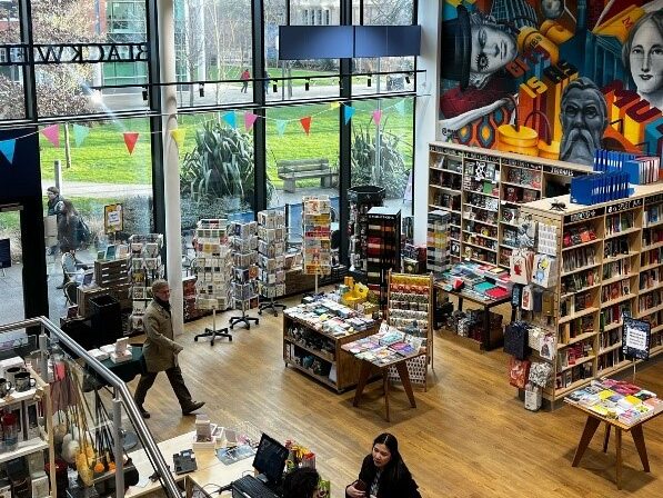 The interior of Blackwells bookshop, taken from the top of a staircase. The camera is angled down to a room with a hardwood floor filled with bookshelves, tables, and postcard racks. The left wall is made entirely of glass, with patches of grass and a pavement visible outside. Customers mill around the room. A large red, blue, and yellow Brutalist mural is visible on the upper right wall.