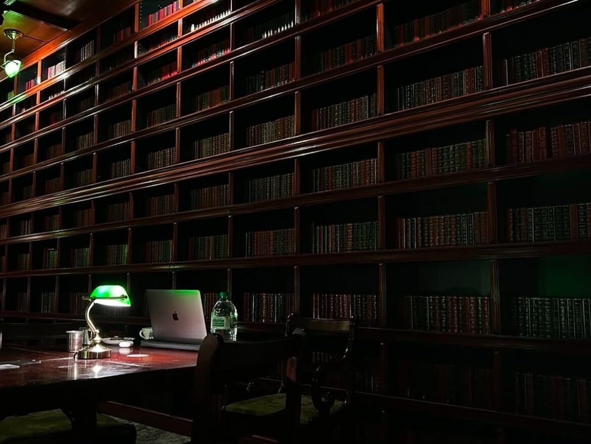 the library inside Mala bookshop and bar. A computer and water bottle are lit up by a green librarian's lamp. The rest of the room is fark, with tall, dark bookshelves stretching from floor to ceiling behind.
