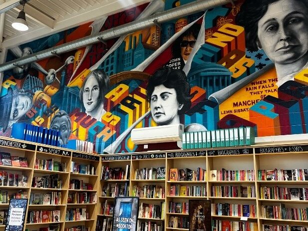 The interior of Blackwells bookshop. In the bottom half of the image are rows of bookshelves. Above them is a Brutalist mural painted in red, blue, and yellow, with greyscale portraits of various women. 