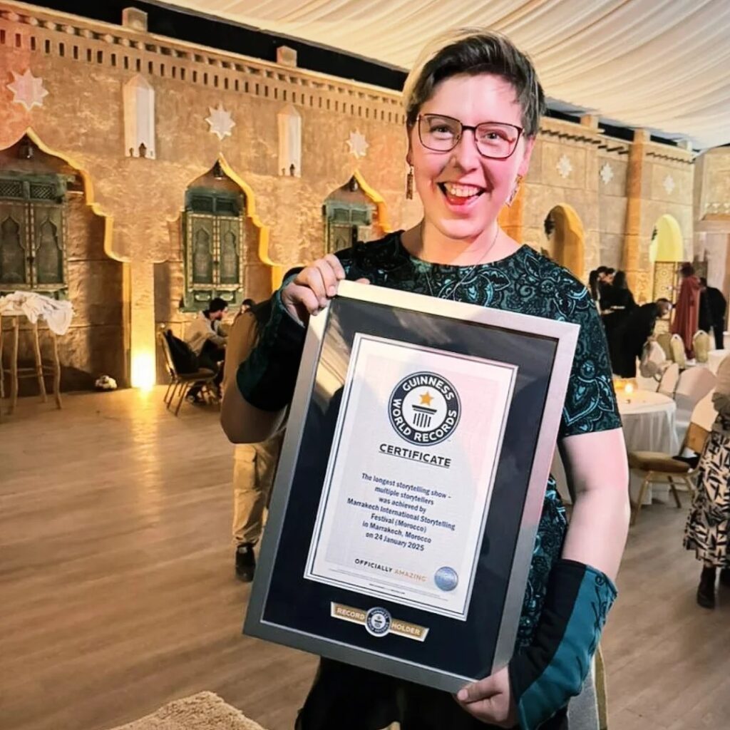 Iris Skipworth, a white woman with short blinde hair, red glasses, and a green dress and gloves, stands smiling at the camera holding a framed certificate. The certificate has a silver frame and says 'Guinness World Records Certificate: The longest storytelling show - multiple storytellers - was achieved by Marrakech International Storytelling Festival (Morocco) in Marrakech Morocco on 24 January 2025'. She is standing in a room of tables and chairs, with stars on the wall and a cloth ceiling, lit up by lamps.