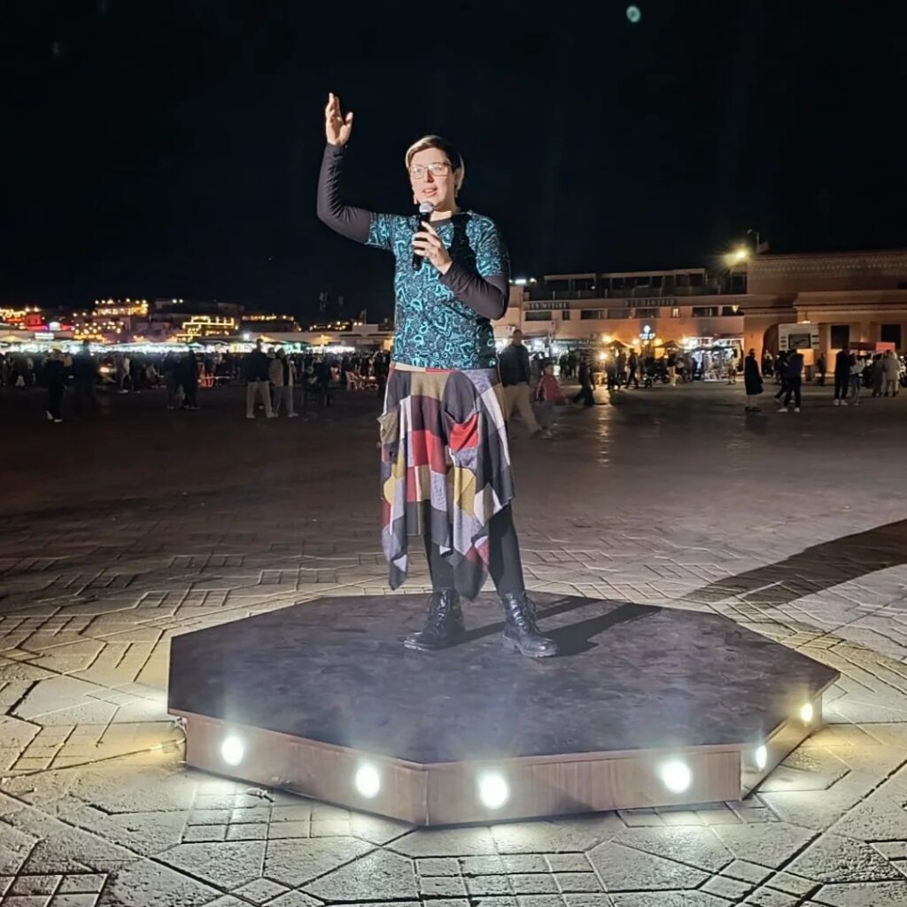Iris Skipworth, a white woman with glasses and short blonde hair, stands on an octagonal stage fitted with lights. She is wearing a blue shirt and a patchwork skirt with boots, and holds a microphone. She has her other arm raised, and is smiling at something behind the camera. It is night time.