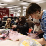 Dr Lynn Setterington, a white woman with short brown hair and a blue checked button-up shirt, leans over a table on the right side of the image. She is sharing the table with a crowd of women, and the table is covered in scraps of cloth, scissors, and other tools. They are hard at work stitching something together in a large, open-plan studio.