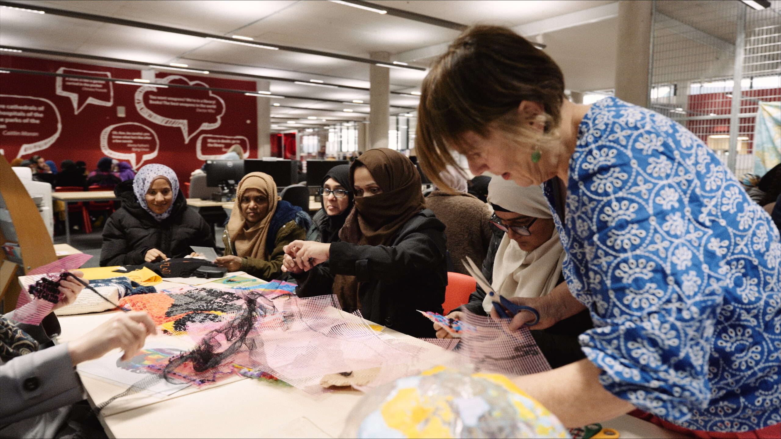 Dr Lynn Setterington, a white woman with short brown hair and a blue checked button-up shirt, leans over a table on the right side of the image. She is sharing the table with a crowd of women, and the table is covered in scraps of cloth, scissors, and other tools. They are hard at work stitching something together in a large, open-plan studio.