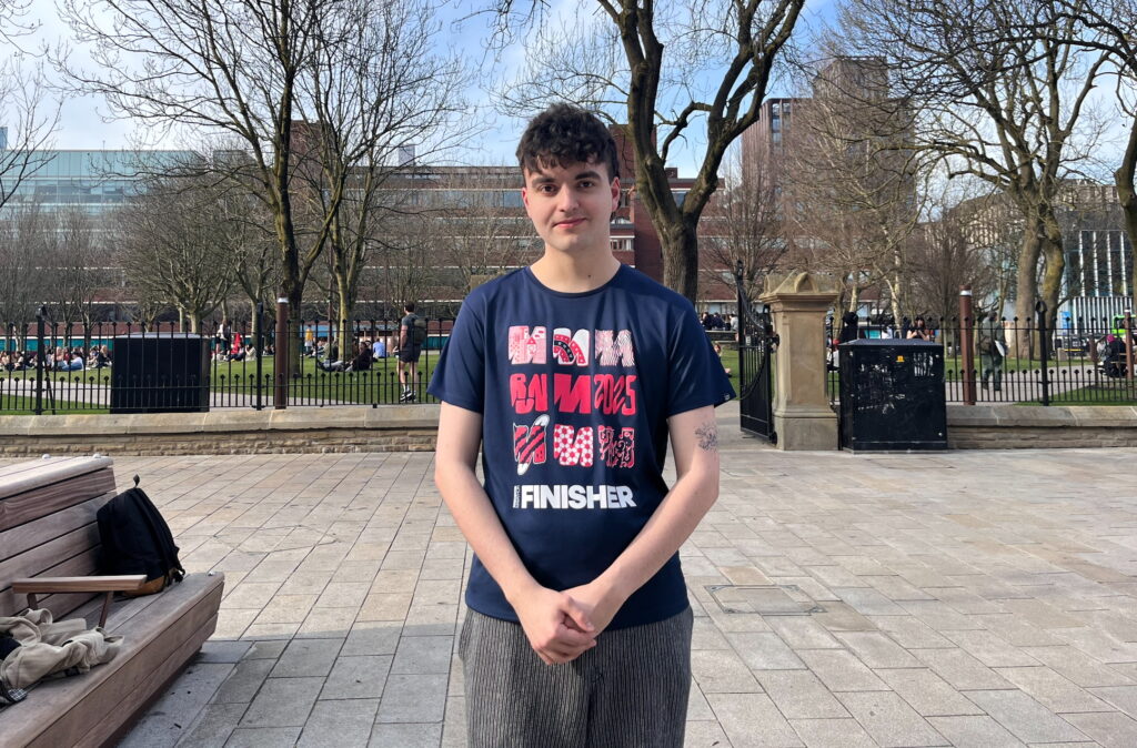 Kian Godbold is a young white man with short brown hair. He is wearing a dark T-shirt with three rows of red and pink 'M's on it. He has his hands folded on his front, and is standing on a pavement outside a park.