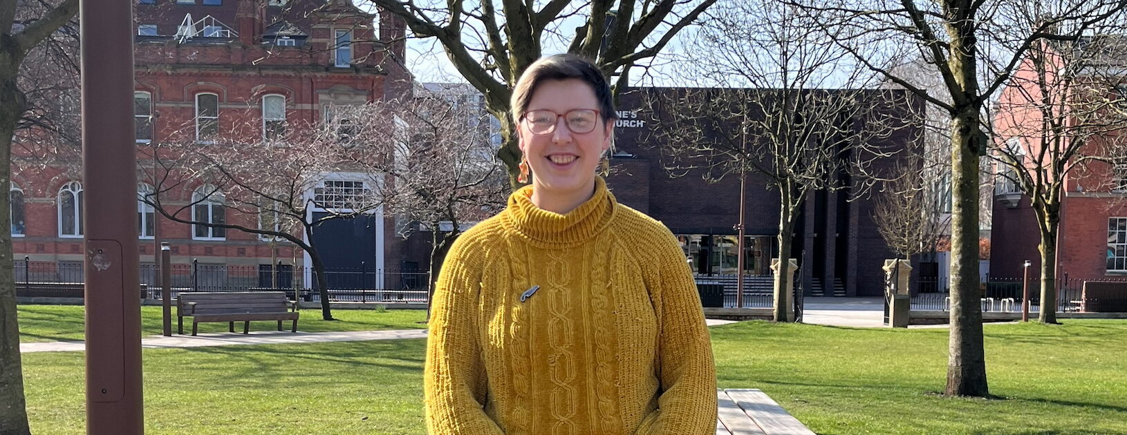 Iris Skipworth, a white woman with short blonde hair, glasses, yellow jumper, and blue jeans, stands with her legs crossed and hands clasped, smiling a the camera. She is leaning against a picnic table in All Saints Park, with trees and grass in the background. It is a sunny day.