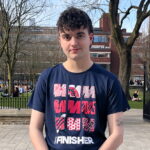 Kian Godbold is a young white man with short brown hair. He is wearing a dark T-shirt with three rows of red and pink 'M's on it. He has his hands folded on his front, and is standing on a pavement outside a park.