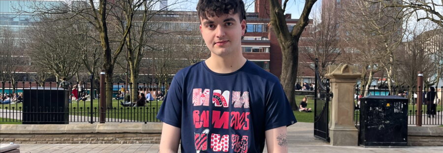 Kian Godbold is a young white man with short brown hair. He is wearing a dark T-shirt with three rows of red and pink 'M's on it. He has his hands folded on his front, and is standing on a pavement outside a park.