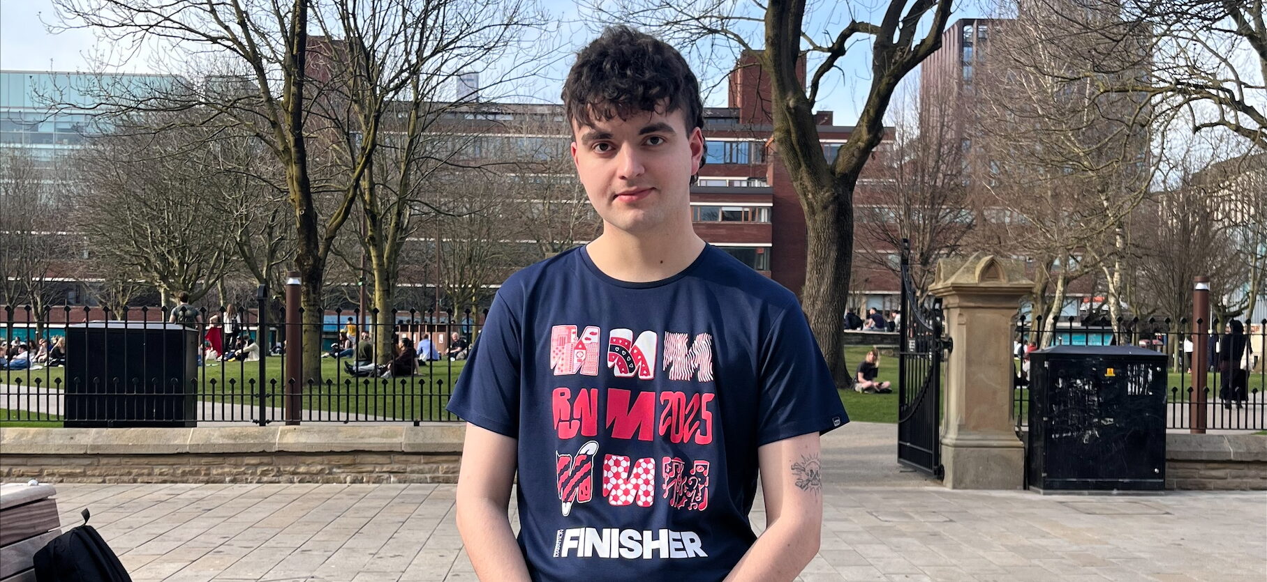 Kian Godbold is a young white man with short brown hair. He is wearing a dark T-shirt with three rows of red and pink 'M's on it. He has his hands folded on his front, and is standing on a pavement outside a park.