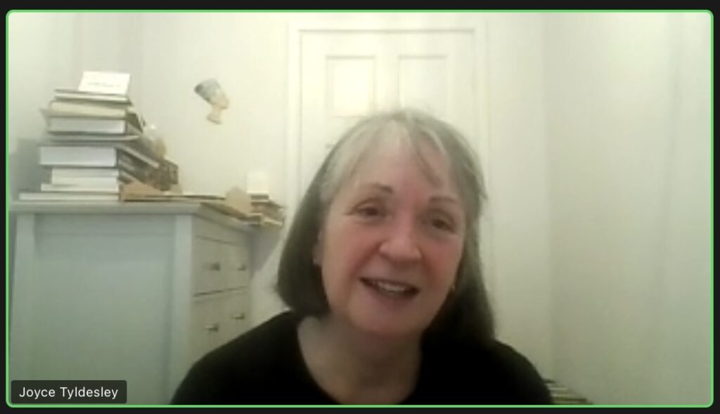 Joyce Tyldesley, a white woman with long grey hair and a fringe, is smiling mid-speech while on a Zoom call. She sits in a white room with a cabinet behind her, piled with books on the top. 