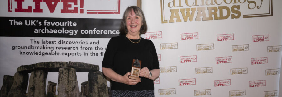 Joyce Tyldesley, a white woman with long grey hair and fringe, stands in a black dress holding a trophy, smiling at the camera. She is standing in front of two signs, which read: 'CURRENT ARCHAEOLOGY LIVE' and 'CURRENT ARCHAEOLOGY AWARDS'