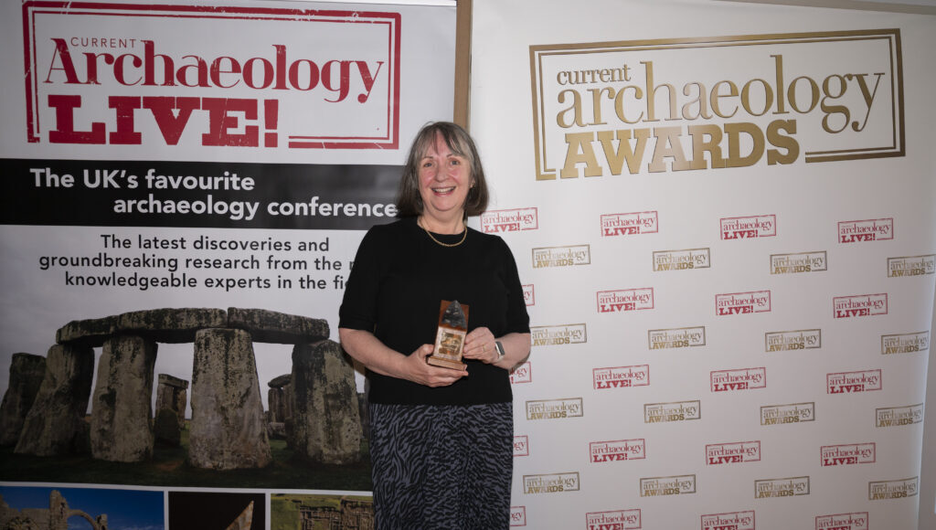 Joyce Tyldesley, a white woman with long grey hair and fringe, stands in a black dress holding a trophy, smiling at the camera. She is standing in front of two signs, which read: 'CURRENT ARCHAEOLOGY LIVE' and 'CURRENT ARCHAEOLOGY AWARDS'