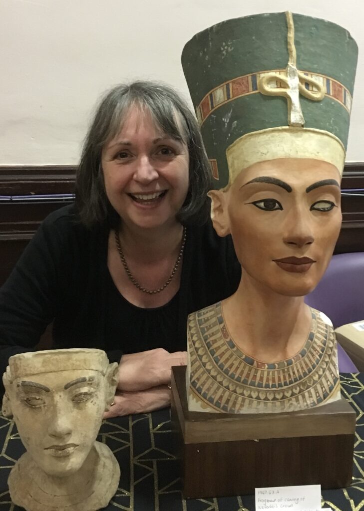 Joyce Tyldesley, a white woman with long grey hair and a fringe, grins into the camera while leaning across a table. In front of her on either side are two statue busts. On the left of the image is a smaller head with tan skin and no hair. On the right of the image is the bust of Nefertiti, with tan skin, a large turquoise hat adorned with gold, and strings of necklaces.