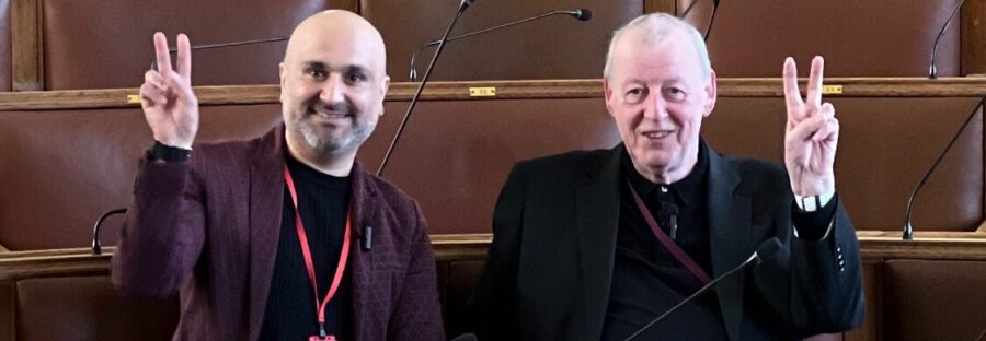 Faraz Arian, a brown man with a bald head and a beard, wears a black shirt and burgundy jacket with a red lanyard. On the right of the image is Cllr John Hacking, an older white man with short white hair, a dark shirt, and a dark suit jacket. Both are smiling at the camera and doing peace signs. They are sat in a council chamber.