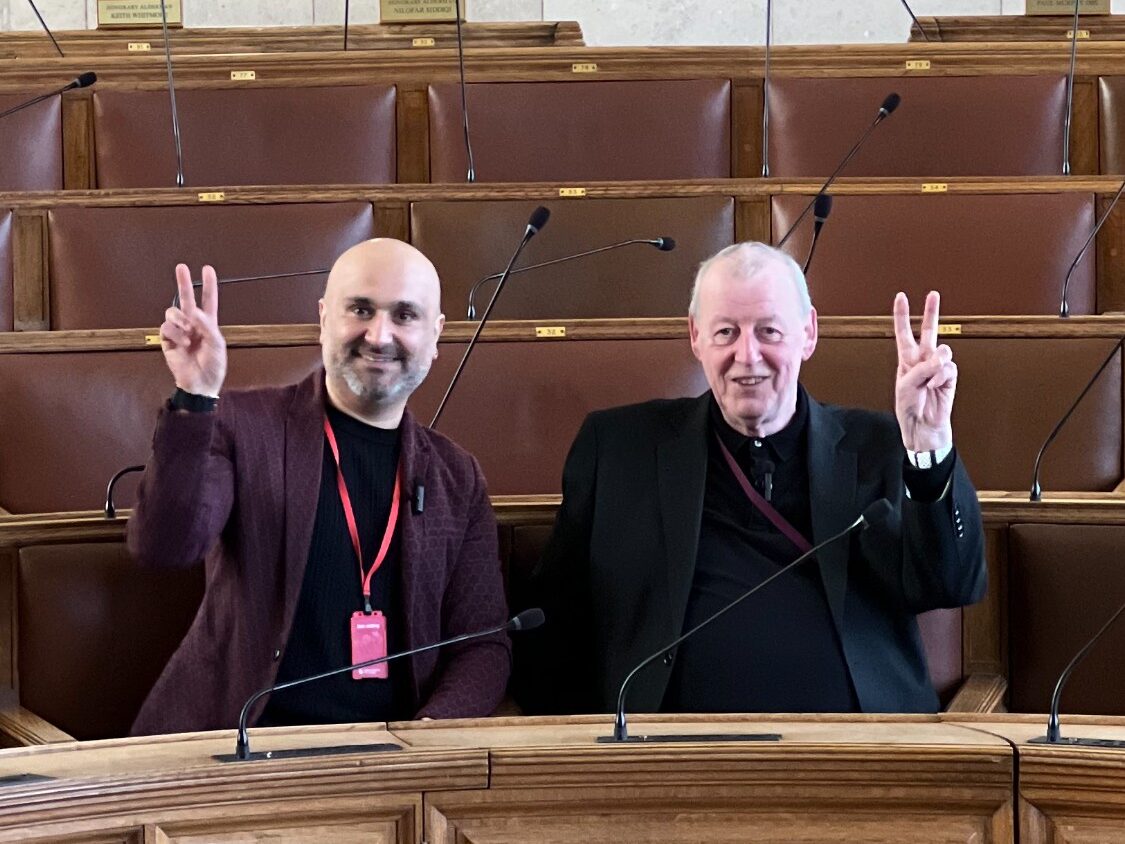 Faraz Arian, a brown man with a bald head and a beard, wears a black shirt and burgundy jacket with a red lanyard. On the right of the image is Cllr John Hacking, an older white man with short white hair, a dark shirt, and a dark suit jacket. Both are smiling at the camera and doing peace signs. They are sat in a council chamber.