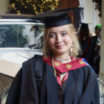Alisson Ponce is a young white woman with wavy, bleach-blonde hair. She is wearing a red and white graduate gown and cap, and standing in front of a vintage car. She is holding a diploma and smiling.