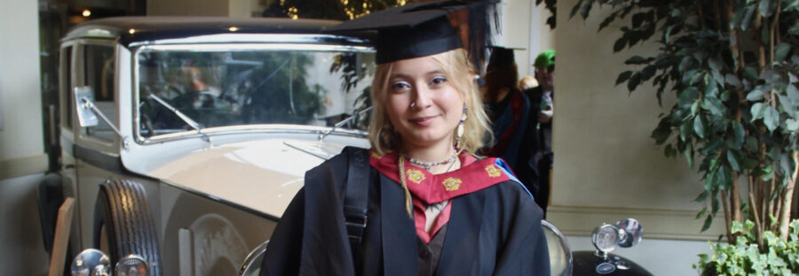 Alisson Ponce feature image Alisson Ponce is a young white woman with wavy, bleach-blonde hair. She is wearing a red and white graduate gown and cap, and standing in front of a vintage car. She is holding a diploma and smiling.