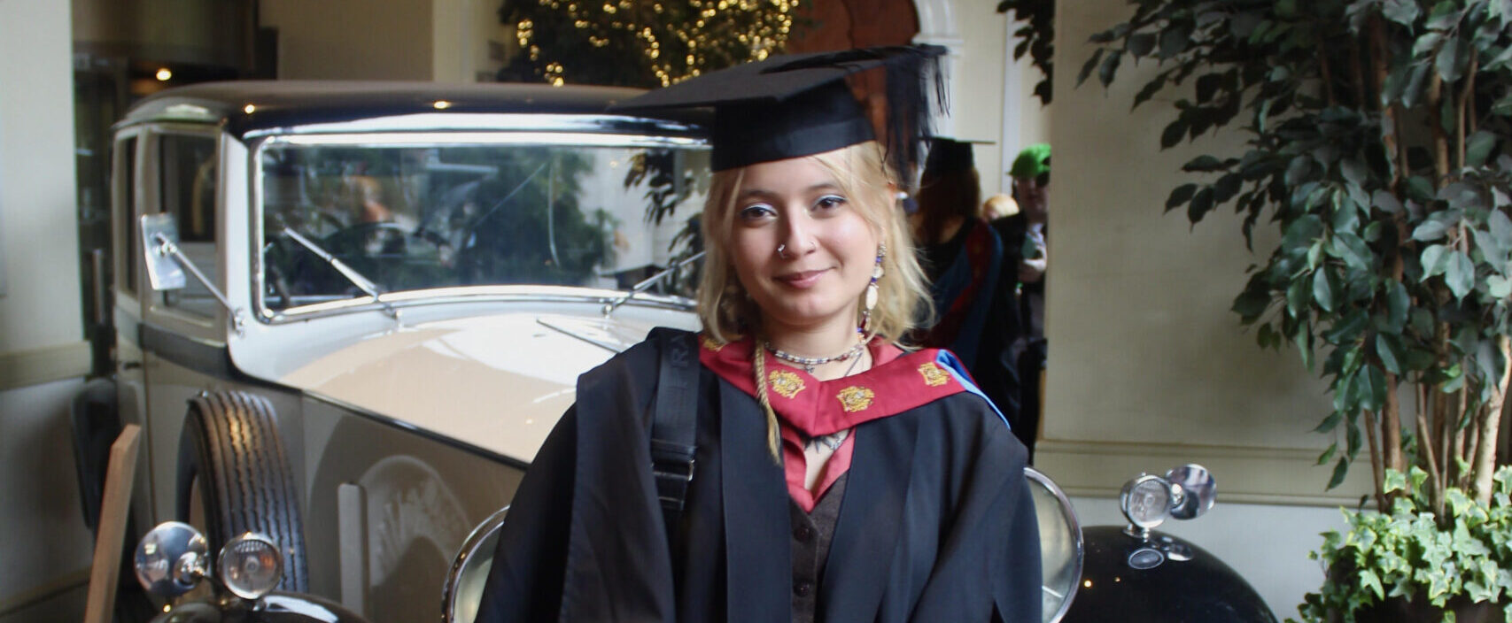 Alisson Ponce is a young white woman with wavy, bleach-blonde hair. She is wearing a red and white graduate gown and cap, and standing in front of a vintage car. She is holding a diploma and smiling.