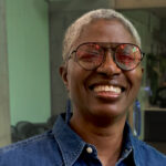 DJ Paulette is a black woman with shaved, grey hair. She is smiling at the camera and wearing pink-tinted sunglasses and a blue denim shirt. She is standing inside a café.