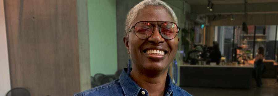 DJ Paulette is a black woman with shaved, grey hair. She is smiling at the camera and wearing pink-tinted sunglasses and a blue denim shirt. She is standing inside a café.