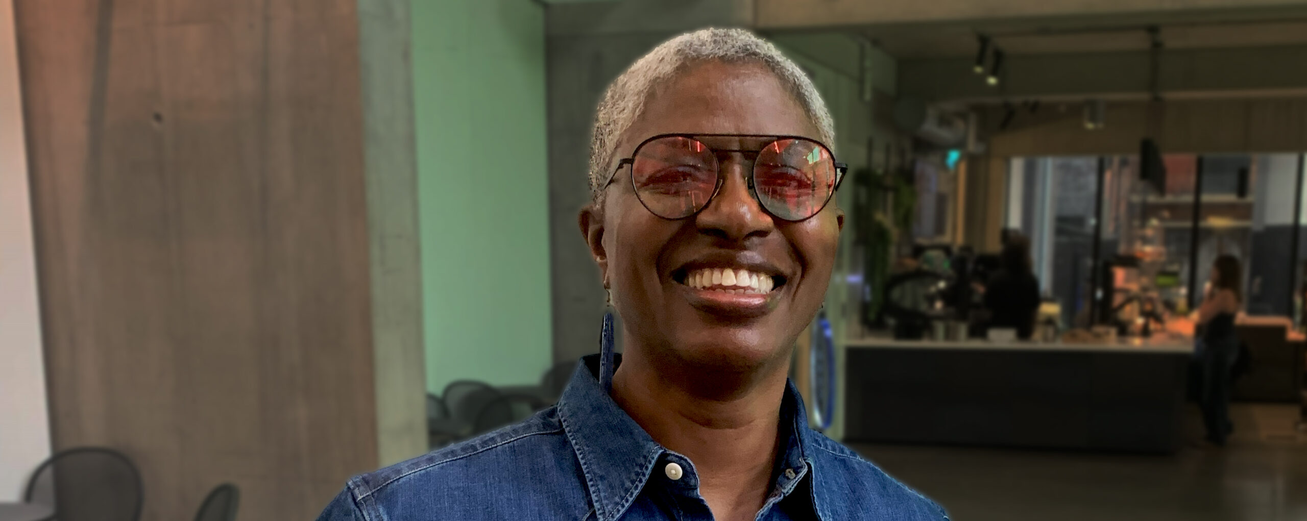 DJ Paulette is a black woman with shaved, grey hair. She is smiling at the camera and wearing pink-tinted sunglasses and a blue denim shirt. She is standing inside a café.