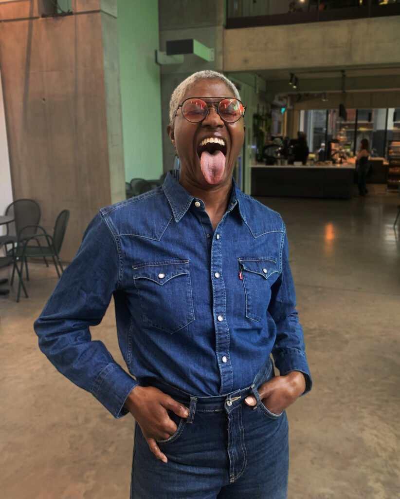 DJ Paulette is a black woman with shaved, grey hair. She is sticking her tongue out at the camera and wearing pink-tinted sunglasses and a blue denim shirt. She is standing inside a café.