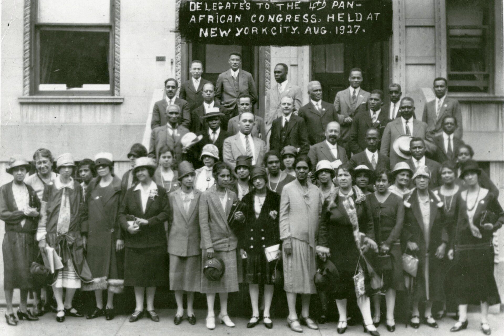 A black and white image of a crowd of people outside a building. The crowd is majority Black, with an even mix of men and women. They are wearing suits or smart dresses typical of 1920s New York. Behind them is a banner which reads: " DELEGATES TO THE 4TH PAN-AFRICAN CONGRESS, HELD AT NEW YORK CITY, AUG 1927"